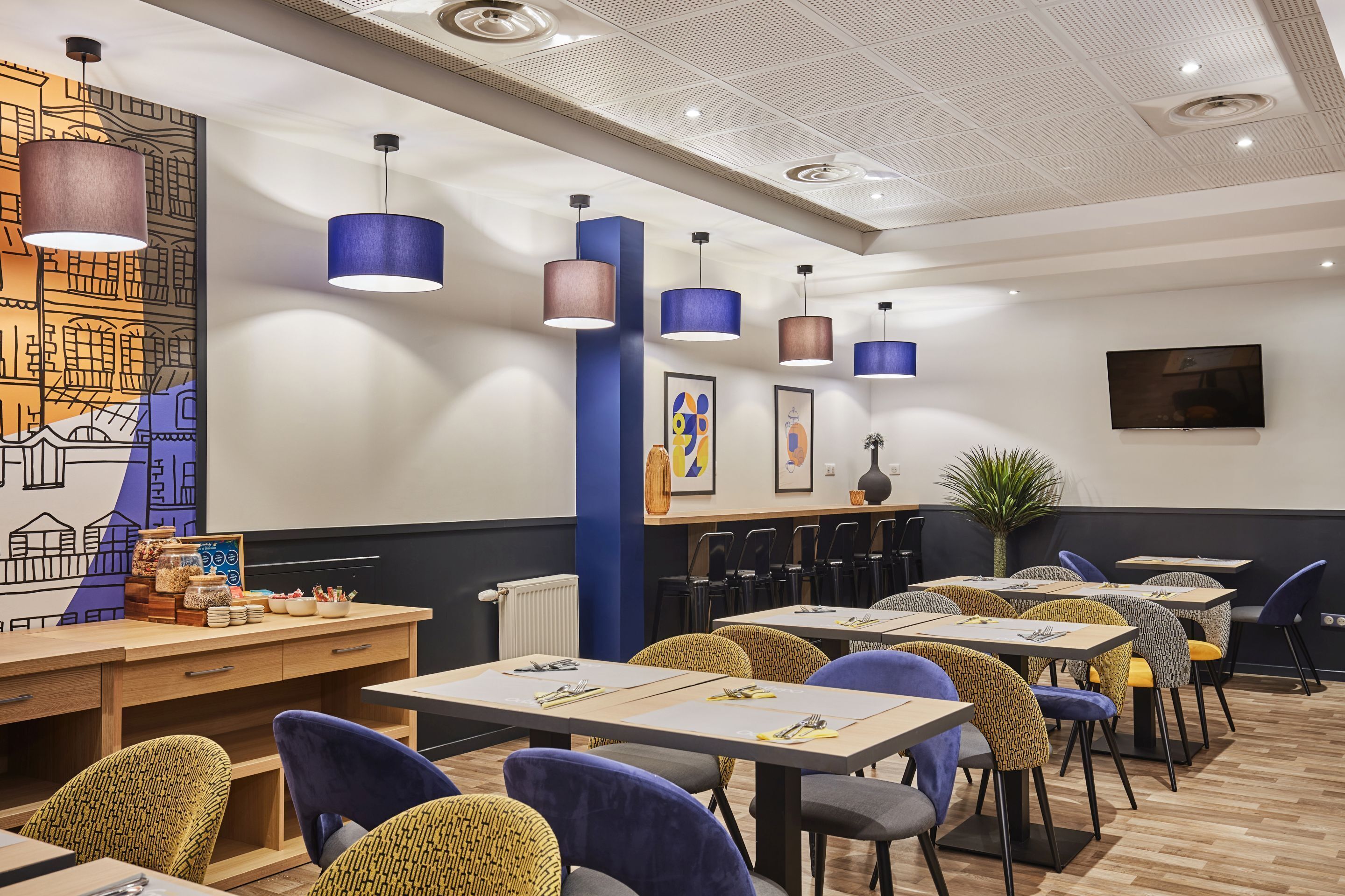 A modern restaurant interior with wooden tables, blue and yellow patterned chairs, and pendant lighting.