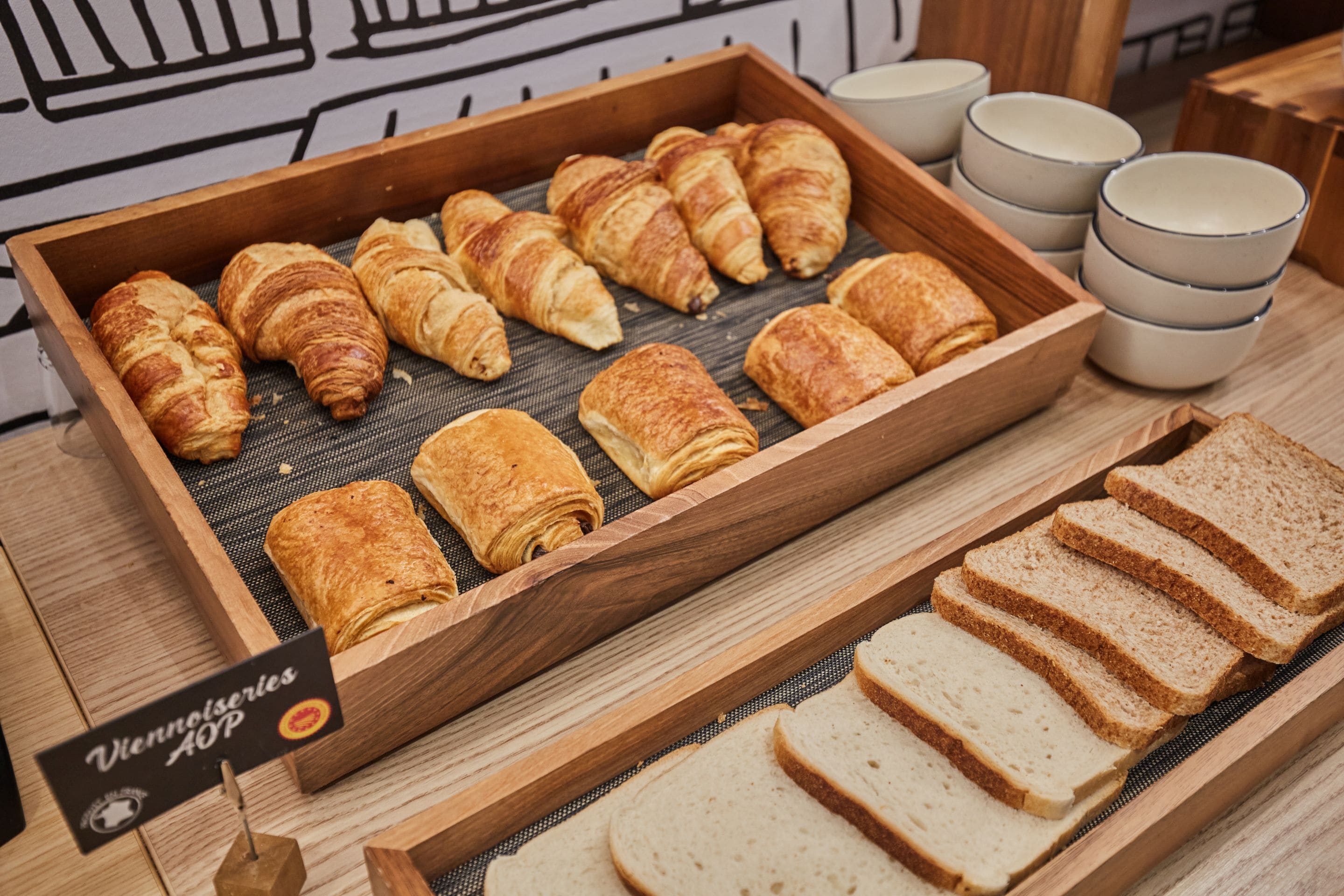 A wooden tray with croissants and sliced bread, accompanied by ceramic cups.