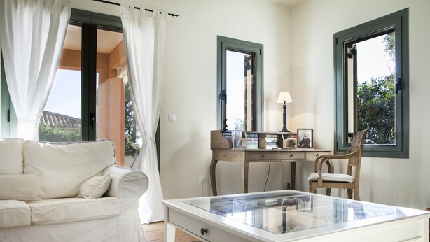 A room with a white sofa, a glass-top table, a wooden desk with a chair, and a window with a view of greenery. - Villa Eleni (Kefalonia)