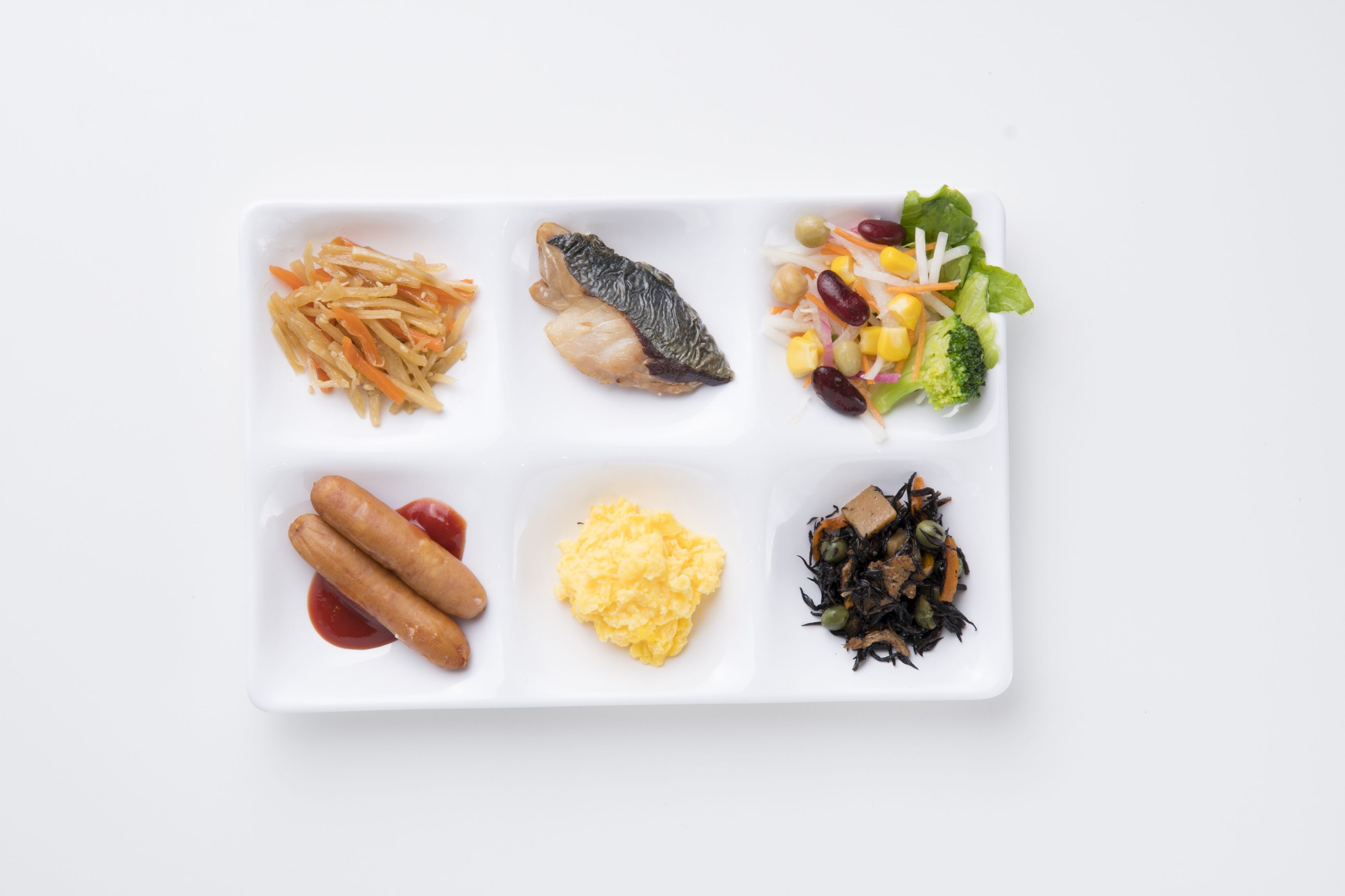 En vit, rektangulär tallrik med sex olika matsorter: strimlad morot, en bit fisk, en sallad med majs, bönor och salladsblad, två korvar med ketchup, äggröra och en liten portion sjögräs.