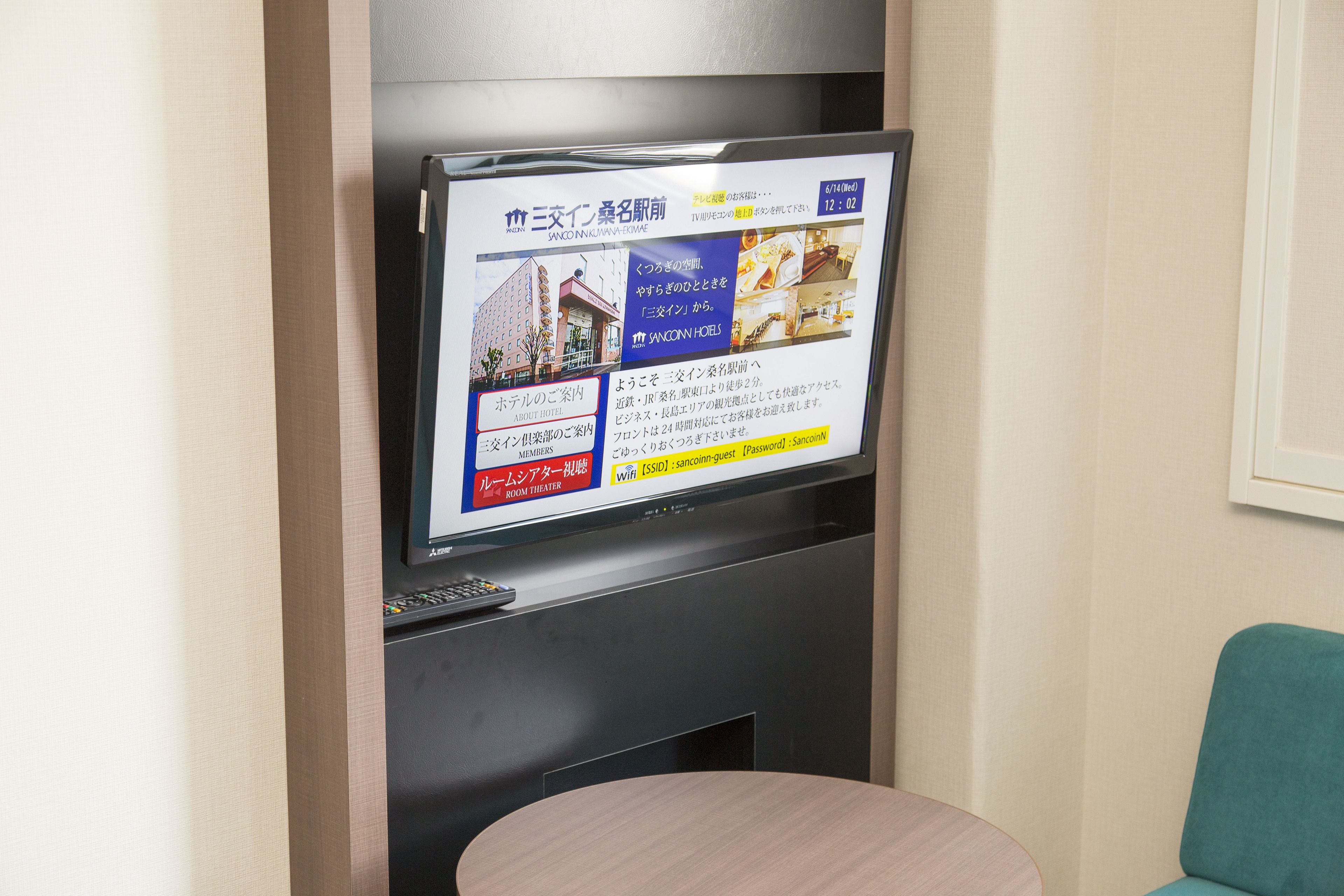 A hotel room with a television displaying a website, a remote control, a small table, and a teal chair.