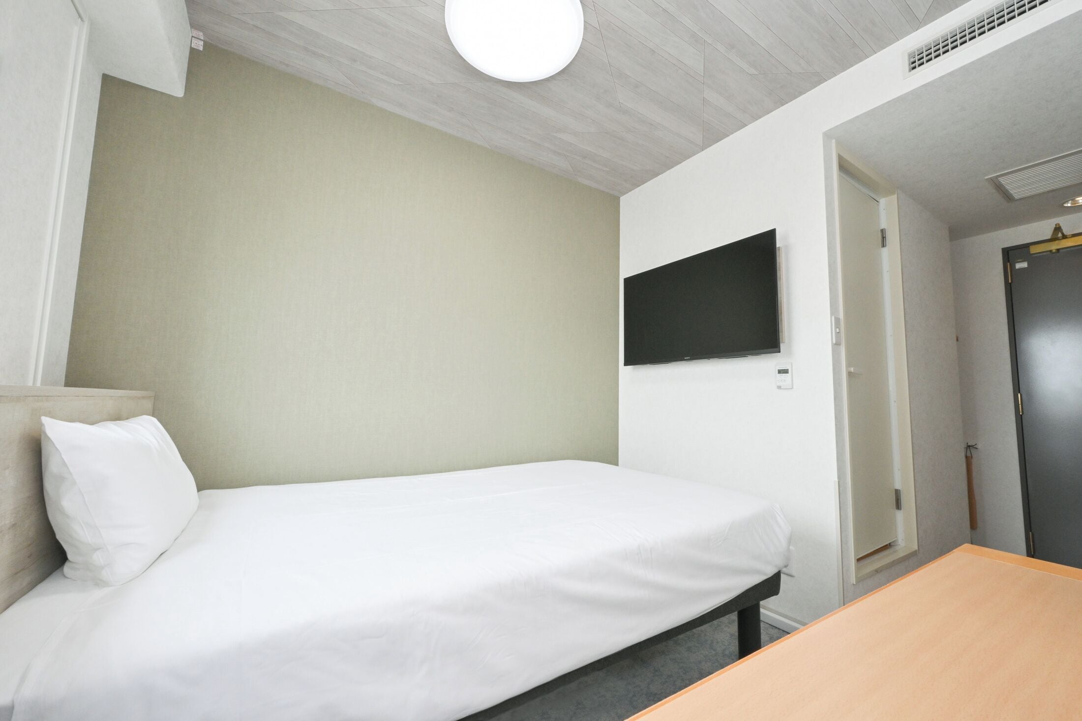 A single bed with a white mattress and pillow, a flat-screen TV mounted on the wall, and a wooden table.