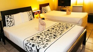 Habitación de hotel con dos camas, cada una con cubrecama y almohadas estampadas. Hay un televisor sobre un mueble bajo y una mesita con una lámpara y una toalla.