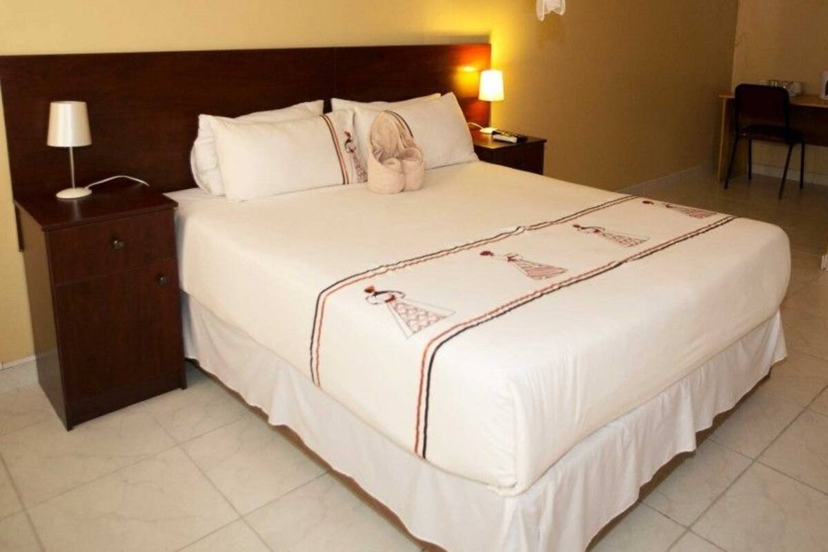 A neatly made bed with a white bedspread and a decorative border, a wooden nightstand with a lamp, and a chair in a small adjoining room.