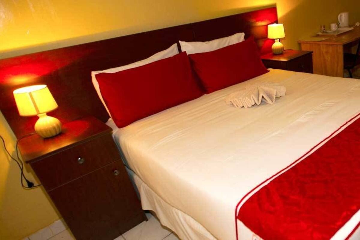 A neatly made bed with red and white bedding, a bedside table with a lamp, and a small desk with a chair.