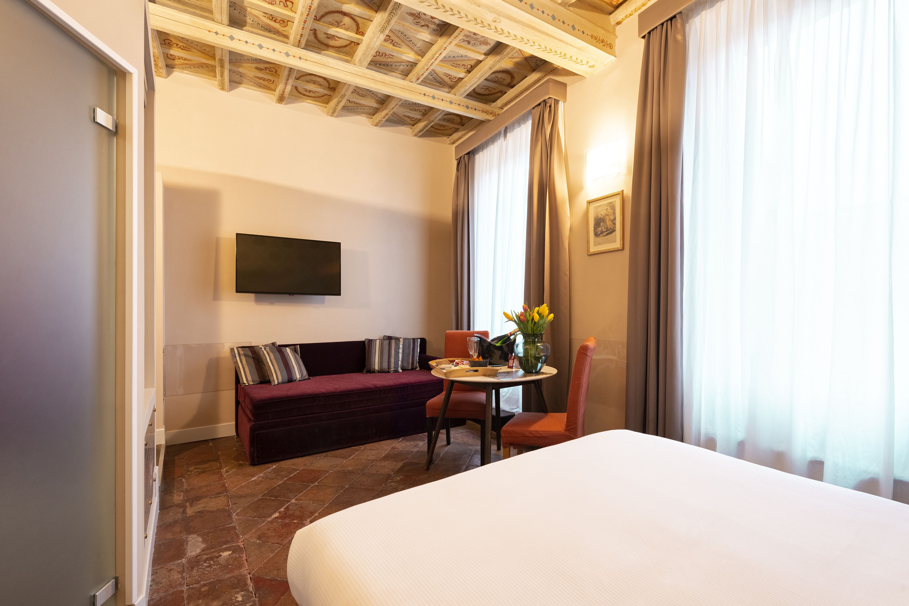 Une chambre d’hôtel avec un lit, un canapé, une table ornée d’un vase de fleurs, un téléviseur fixé au mur et une grande fenêtre avec des rideaux.