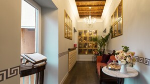 A hallway with a wooden ceiling, a chandelier, a radiator, a sofa, a table with flowers, and a bookshelf. - Le Suite Del Barone (Rome)