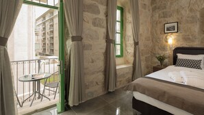 Un dormitorio con pared de piedra, cama con ropa de cama blanca, un pequeño balcón con mesa y silla, y una ventana verde.