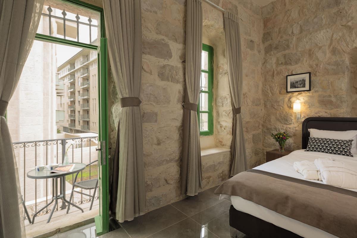 Un dormitorio con pared de piedra, cama con ropa de cama blanca, un pequeño balcón con mesa y silla, y una ventana verde.