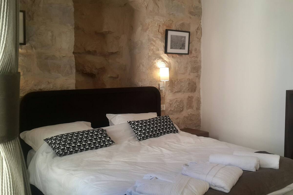 Una cama con ropa de cama blanca y dos almohadas estampadas, una mesita de noche con una lámpara y un cuadro en la pared.