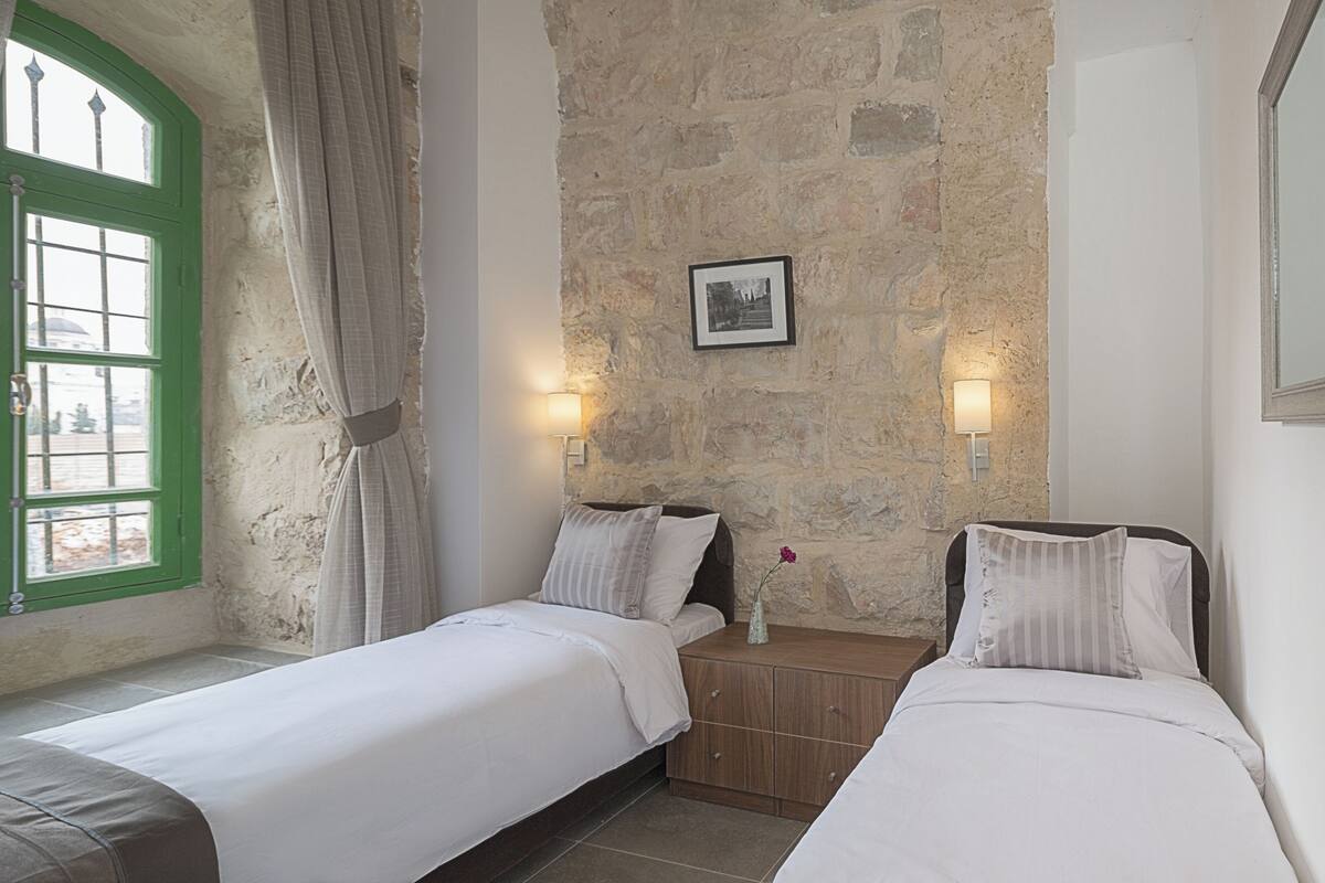 Una habitación con dos camas, una pared de piedra, un cuadro en la pared y una ventana con contraventanas verdes.
