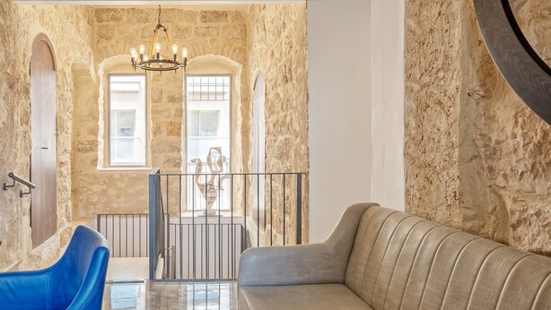Una habitación con paredes de piedra, un candelabro, una silla azul, un sofá color beige y vistas a través de una ventana.