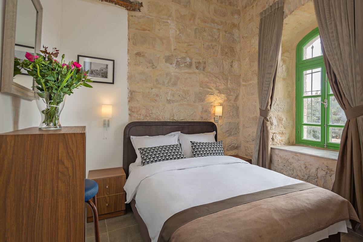 Un dormitorio con pared de piedra, una cama con almohadas estampadas, un escritorio de madera, un jarrón con flores y una ventana con cortinas.