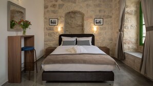 Un dormitorio con pared de piedra, una cama con edredón blanco y marrón, un escritorio de madera con silla azul, y dos luces fijadas a la pared.