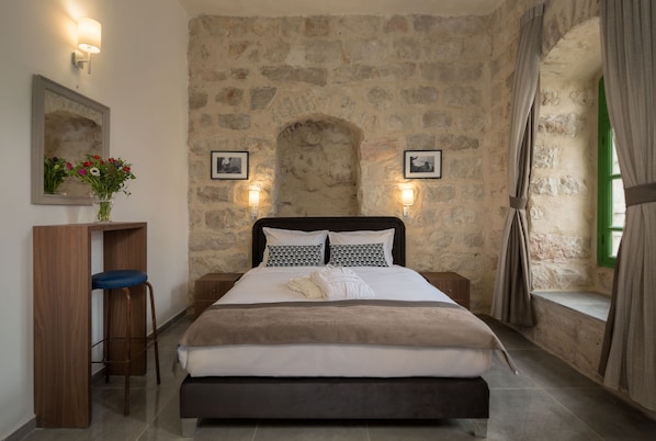 Un dormitorio con pared de piedra, una cama con edredón blanco y marrón, un escritorio de madera con silla azul, y dos luces fijadas a la pared.
