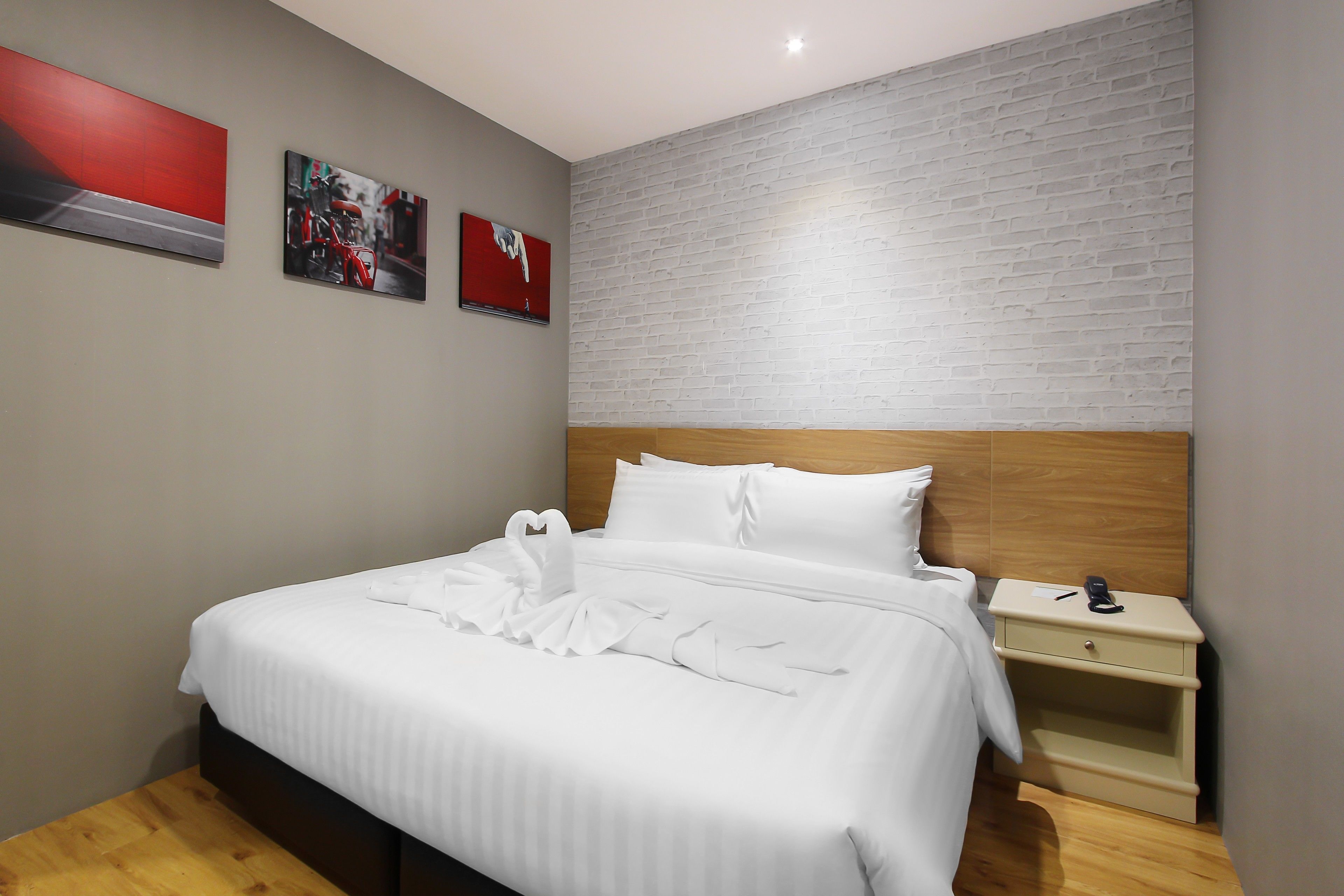 Una cama bien hecha con sábanas blancas y una disposición de toallas con forma de corazón, un cabecero de madera, una mesita de noche con un teléfono y una pared con estampado de ladrillo.