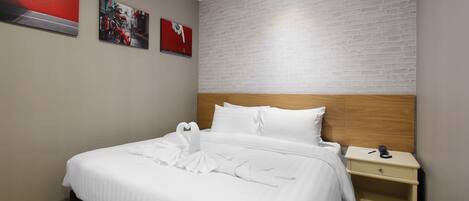 Una cama bien hecha con sábanas blancas y una disposición de toallas con forma de corazón, un cabecero de madera, una mesita de noche con un teléfono y una pared con estampado de ladrillo.