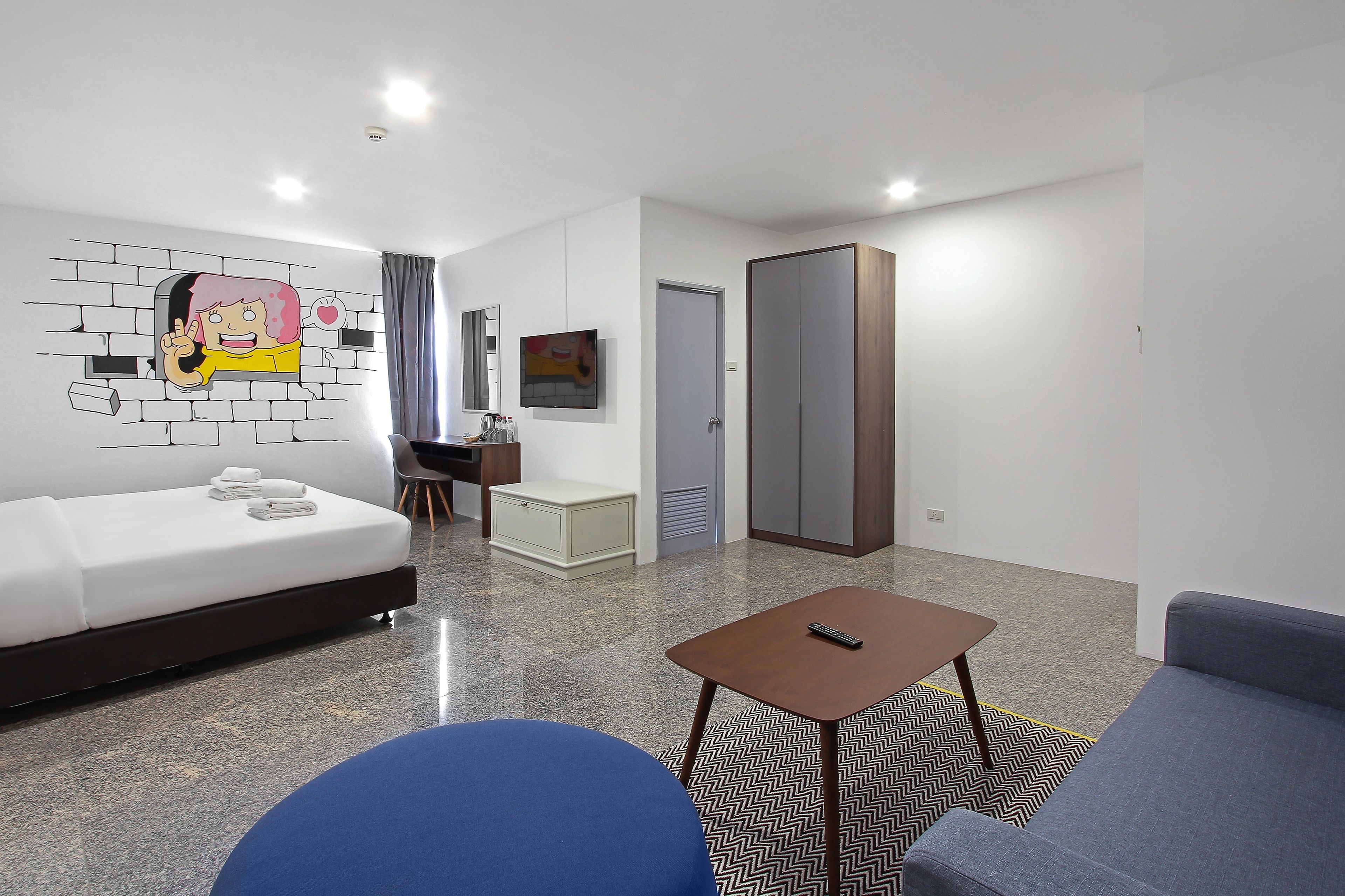 Une chambre d’hôtel moderne avec un mur décoré d’une fresque en briques blanches, un canapé, un fauteuil bleu, une petite table avec une télécommande et un tapis à motifs chevrons.