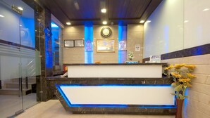 Zona de recepción de un hotel con mostrador de mármol, iluminación LED azul, un reloj, cuadros enmarcados y un jarrón con flores.