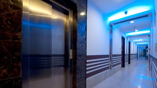 Un pasillo moderno con pared de mármol, ascensor de vidrio y techo iluminado en azul.