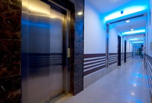 A modern hallway with a marble wall, a glass elevator, and a blue-lit ceiling.