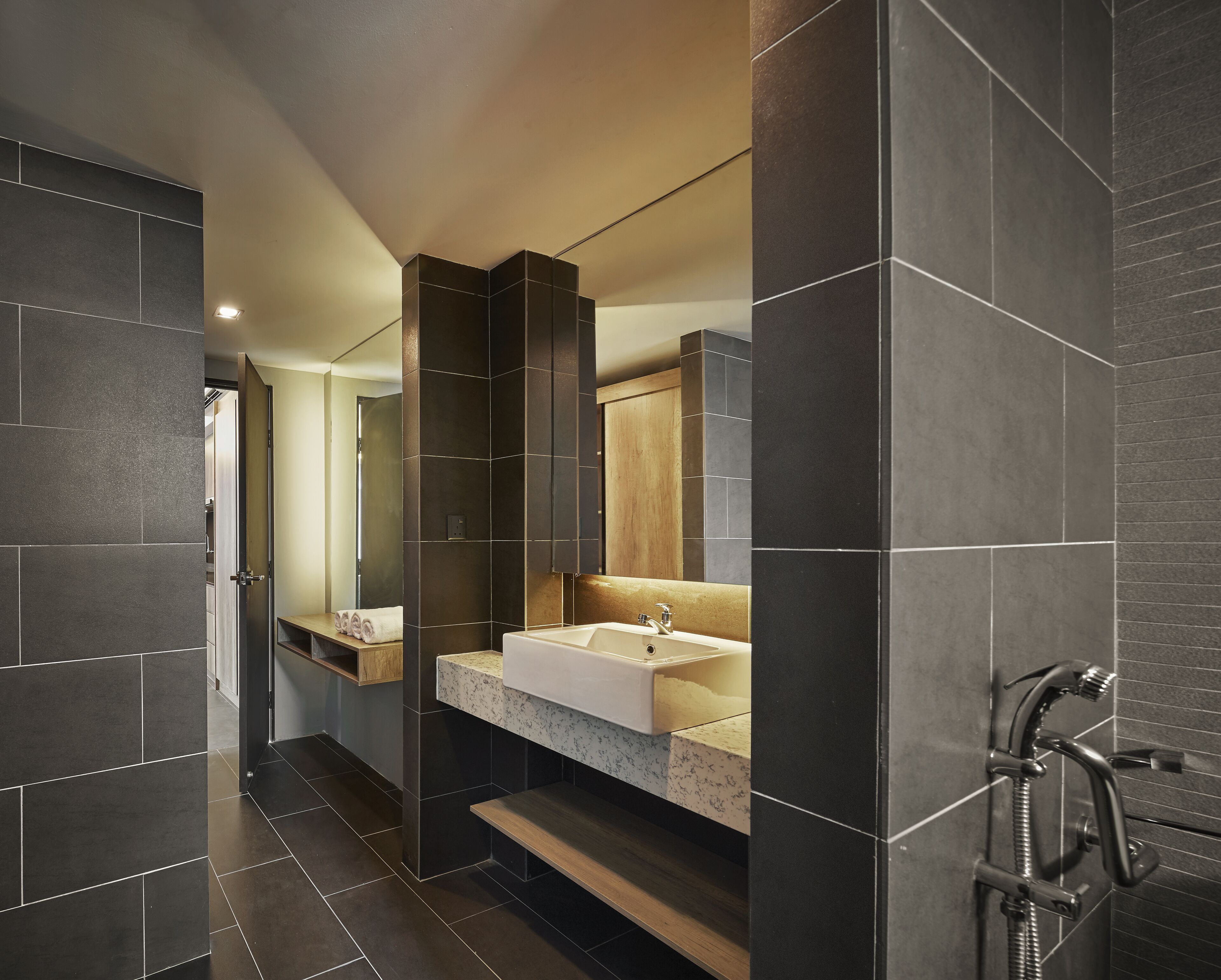 A modern bathroom with dark tiles, a large mirror, a sink with a marble countertop, and a shower area.