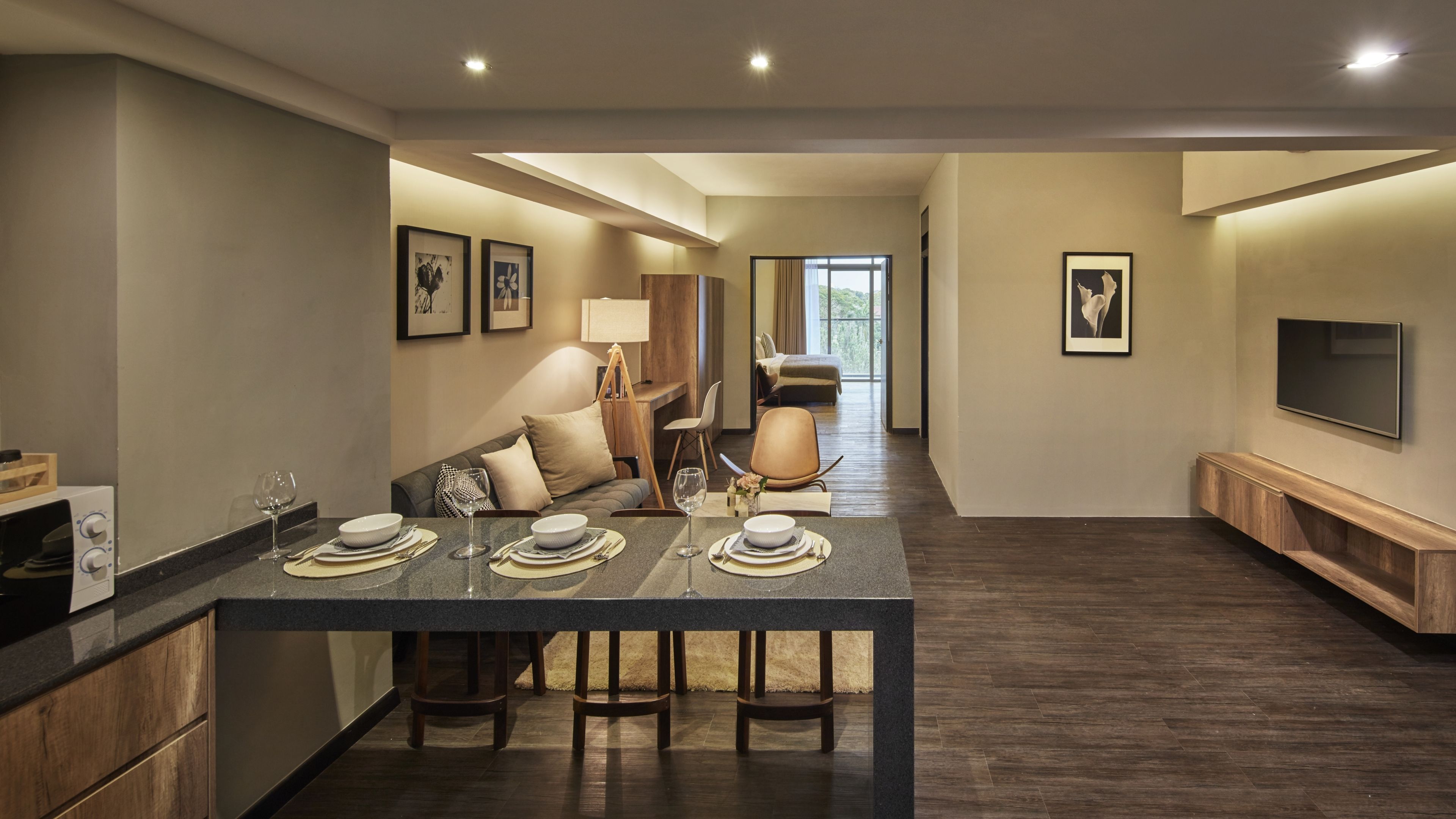A modern hotel dining area with a long table set for four, a sofa, and a TV mounted on the wall.