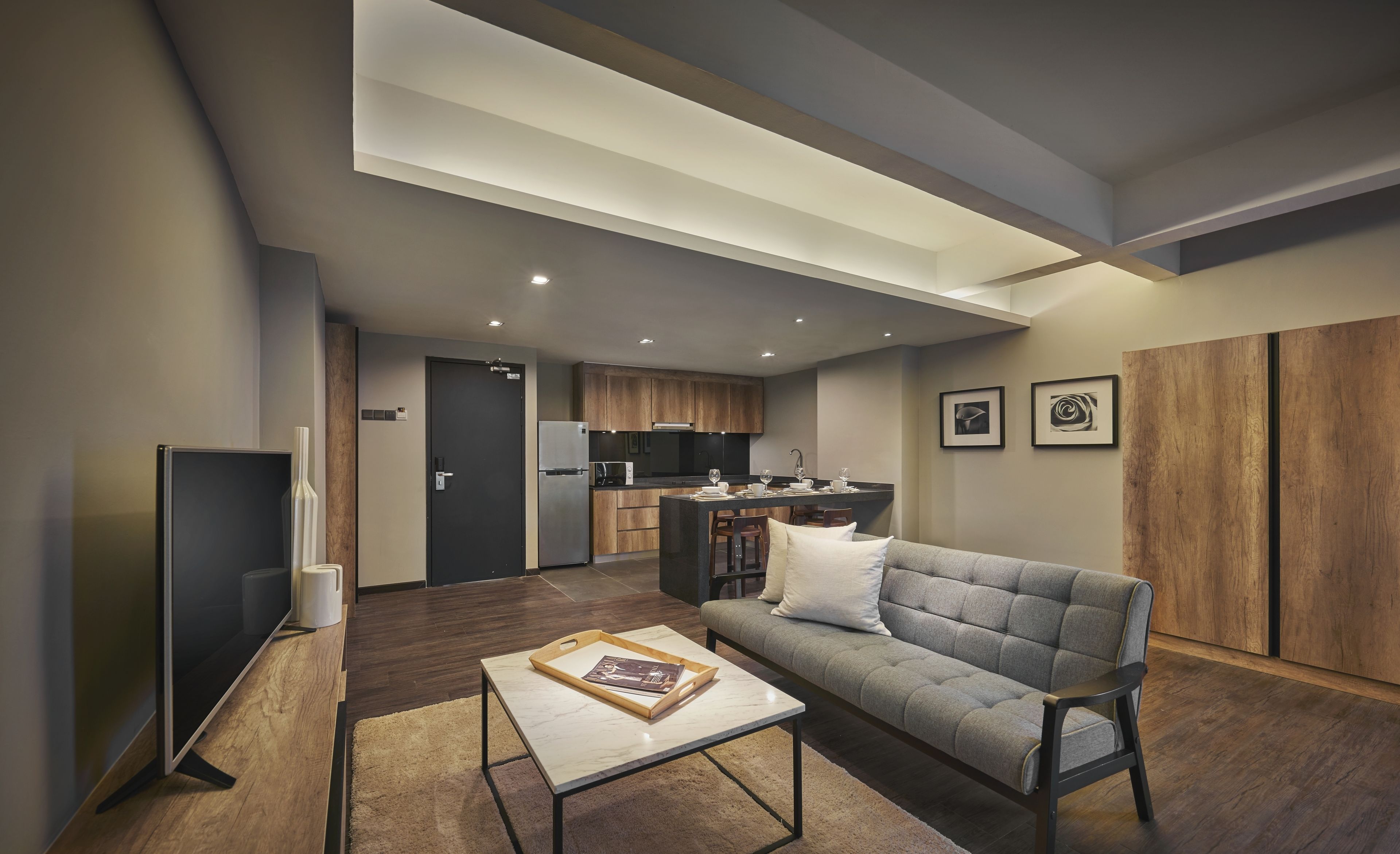 Una sala moderna con un sofá gris, una mesa de centro y un televisor. Hay una zona de cocina con armarios de madera y un refrigerador.