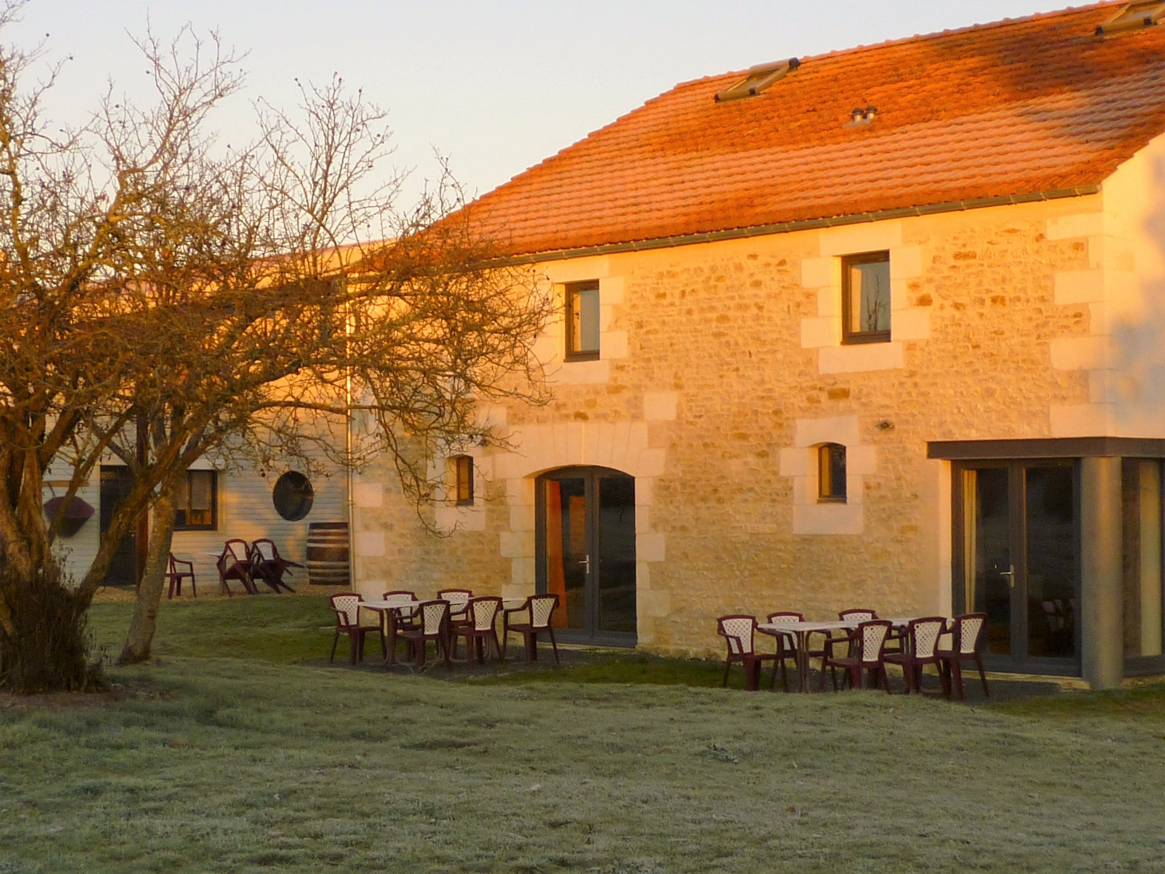A stone building with a tiled roof, outdoor seating area, and a tree in the foreground.