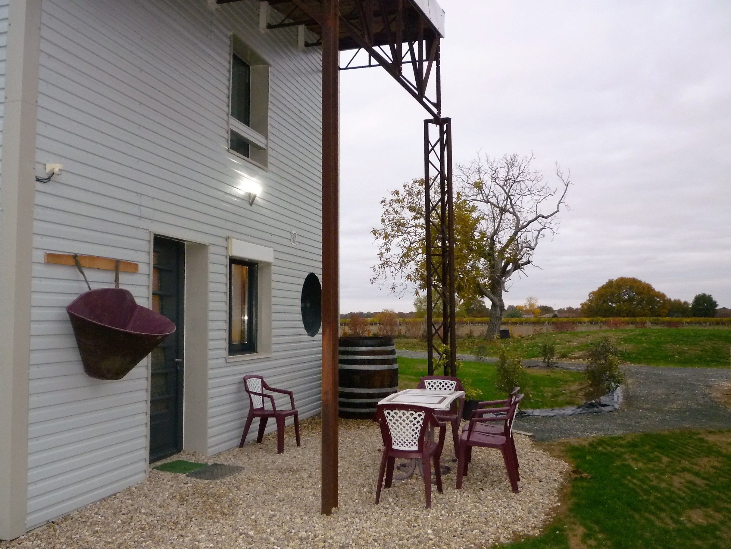 A white building with a covered patio area featuring a table and chairs, a hanging chair, and a small garden with a tree and gravel path.