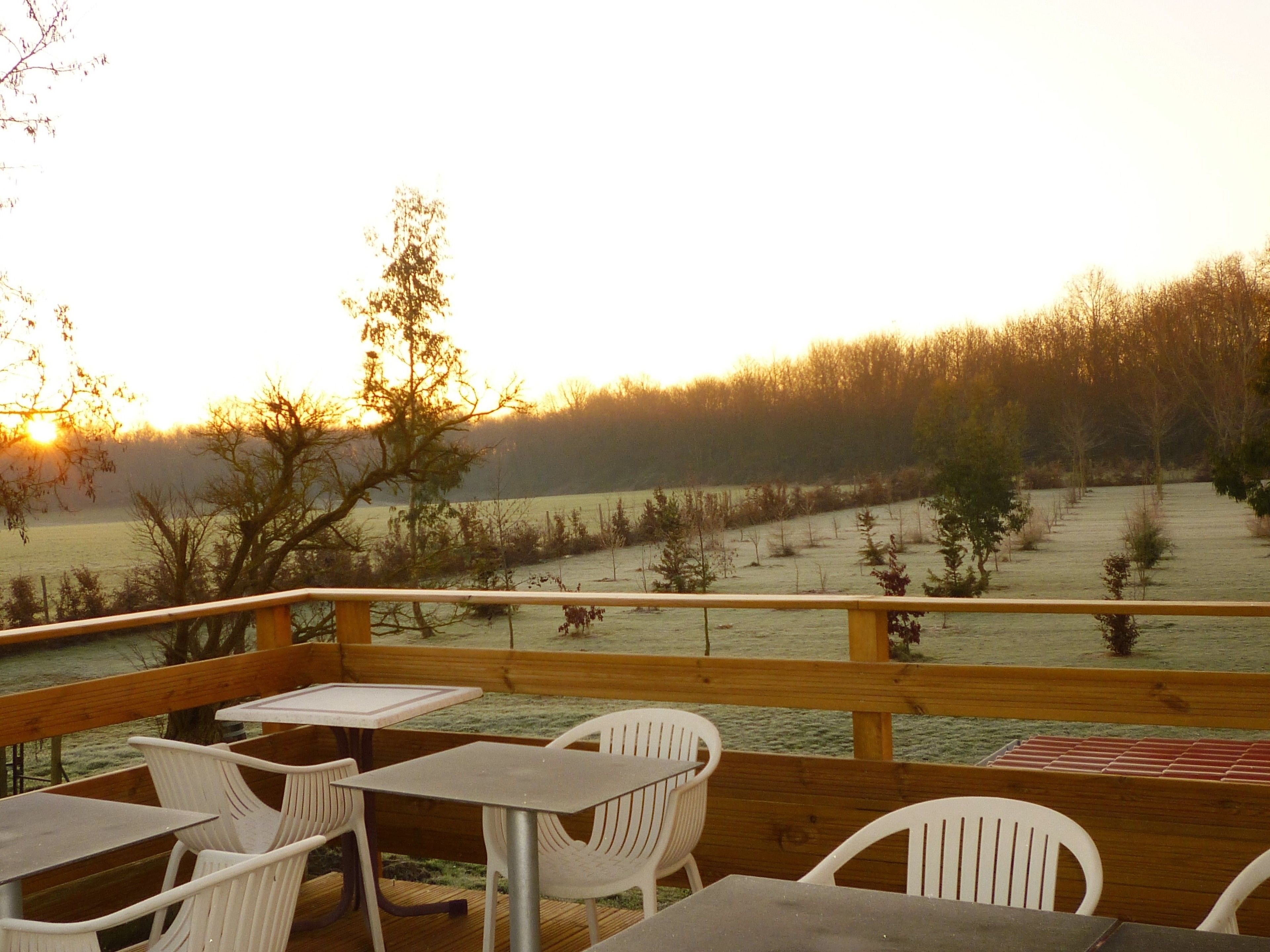 A deck with outdoor seating, overlooking a field and a body of water during sunset.