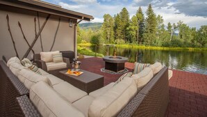 Une terrasse extérieure aménagée avec des meubles en osier, un foyer et une vue sur un lac entouré d’arbres.
