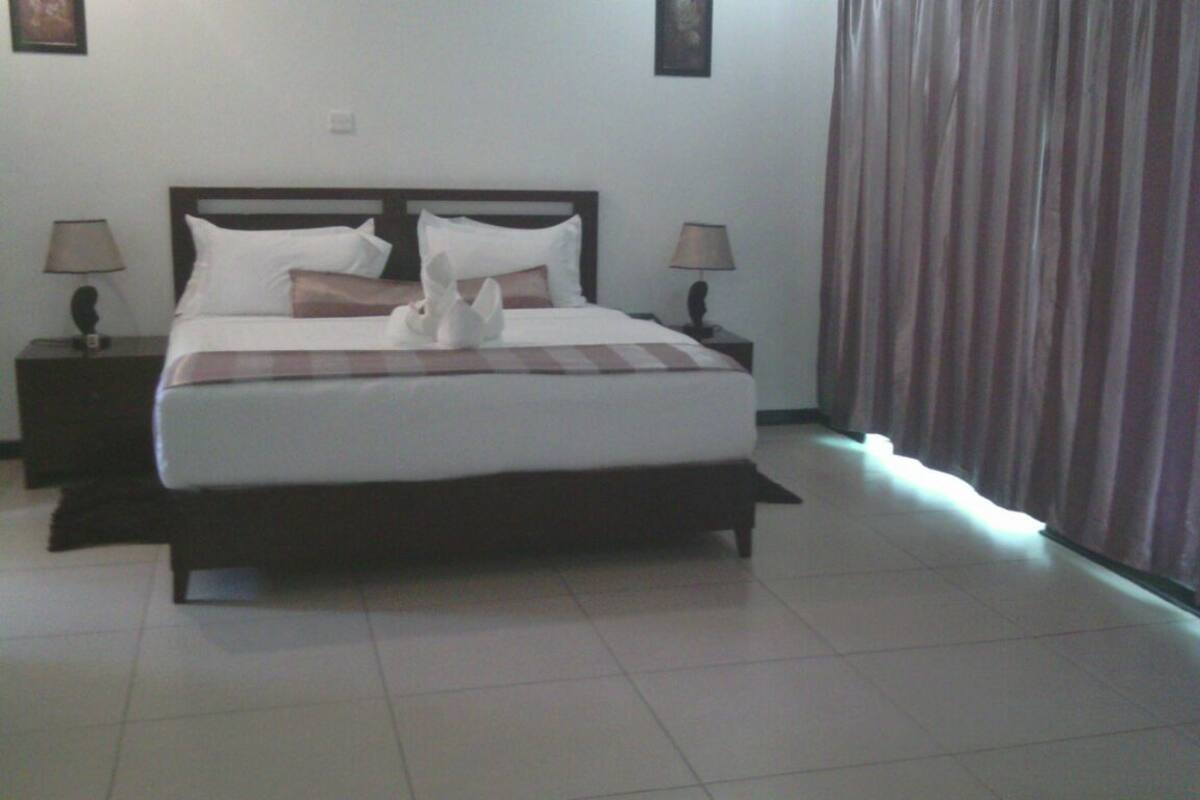 Una cama bien hecha con una manta a cuadros y almohadas blancas, dos mesitas de noche con lámparas, una cortina y un refrigerador.