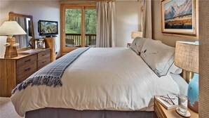 A bedroom with a large bed, wooden dresser, TV, and a balcony with a scenic view.