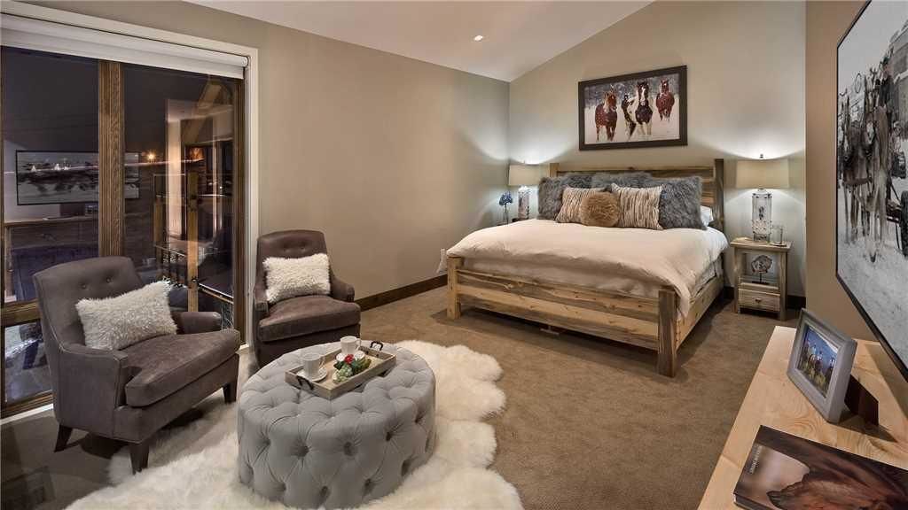 A bedroom with a large bed, two armchairs, a tufted ottoman, a nightstand, and a wall-mounted picture.
