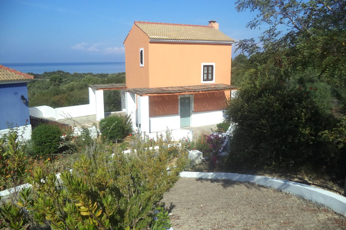 Uma casa de dois andares com telhado em cerâmica, rodeada por vegetação e vista para o mar.