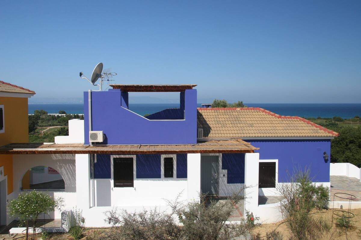 Um edifício com fachada azul e branca, uma antena parabólica e vista para o mar.