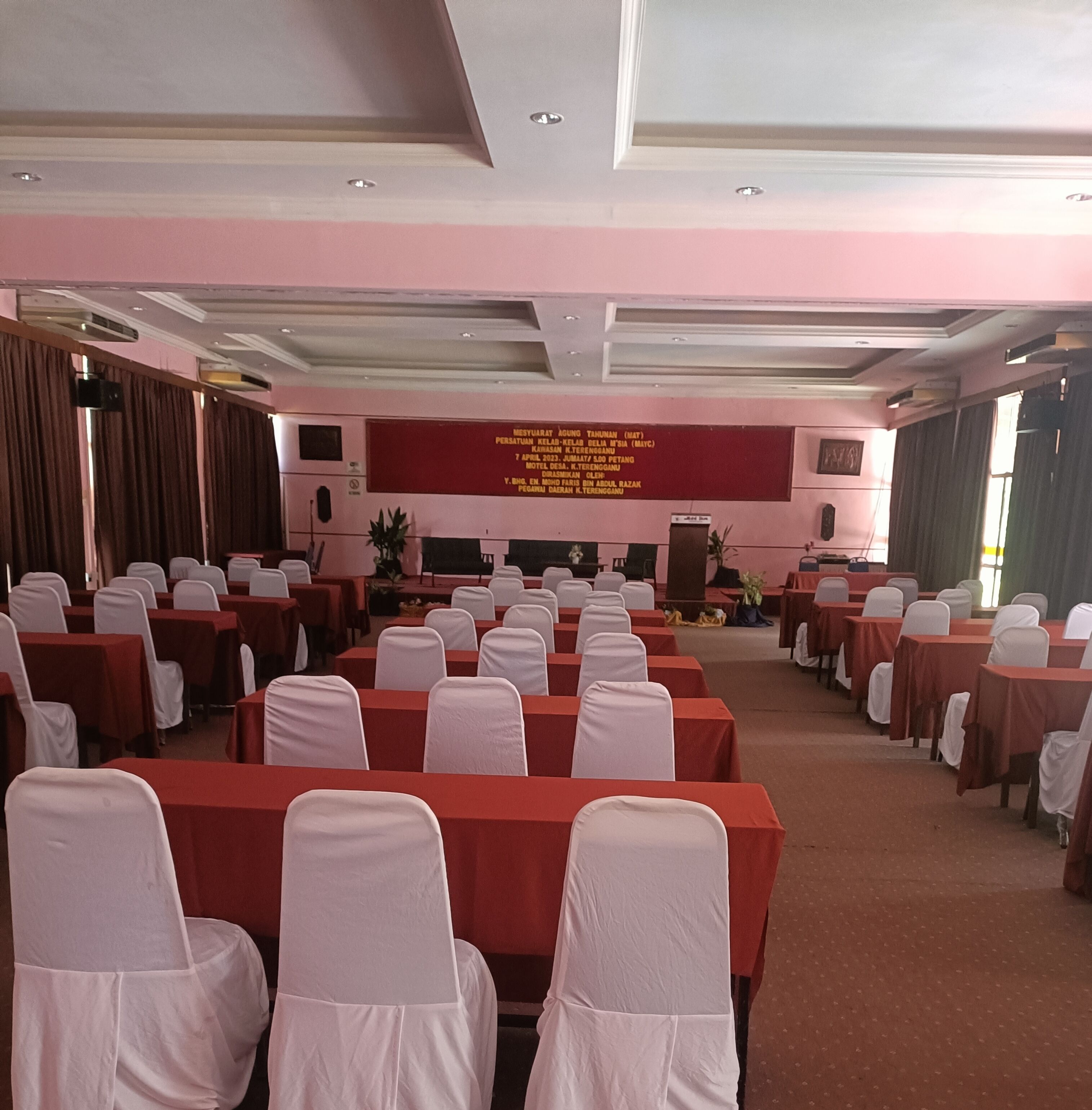 A conference room with rows of tables and chairs, a stage with a podium, and a banner with text.