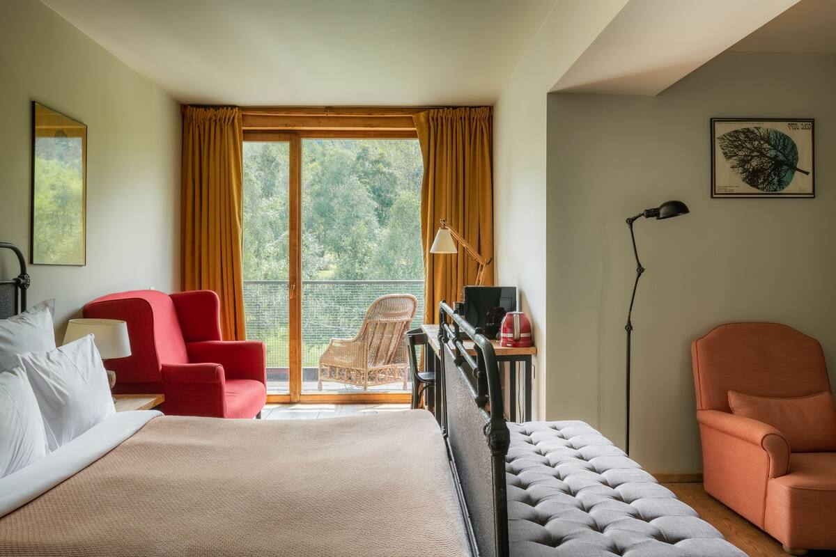 Quarto de hotel com uma cama, duas cadeiras, uma mesa com um computador, uma luminária e vista para o verde pela janela.