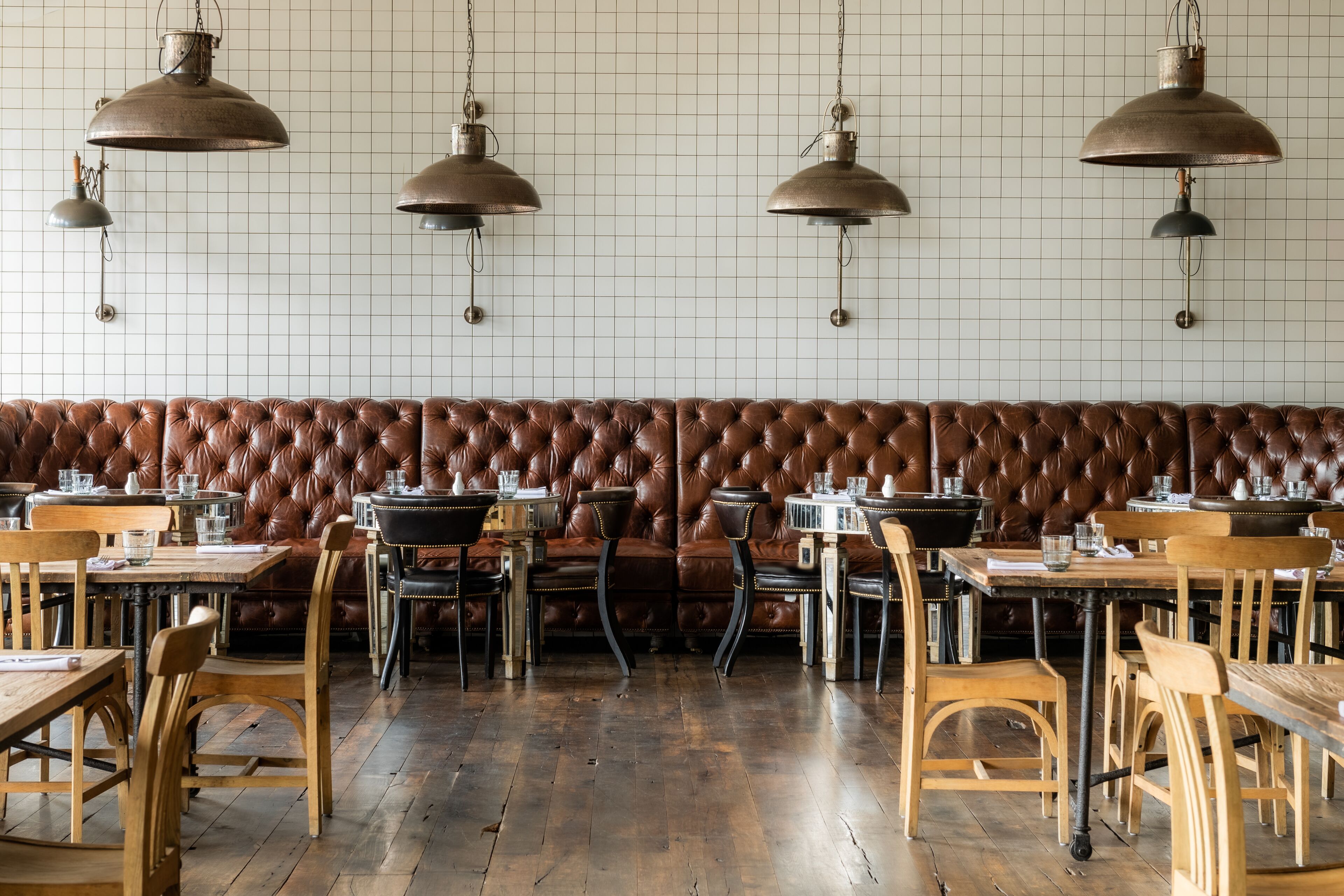 A restaurant interior with a long brown leather booth, wooden tables, and hanging metal lamps.