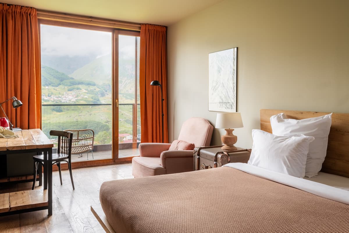 Quarto de hotel com cama, escrivaninha, cadeira, luminária e vista para as montanhas pela janela.