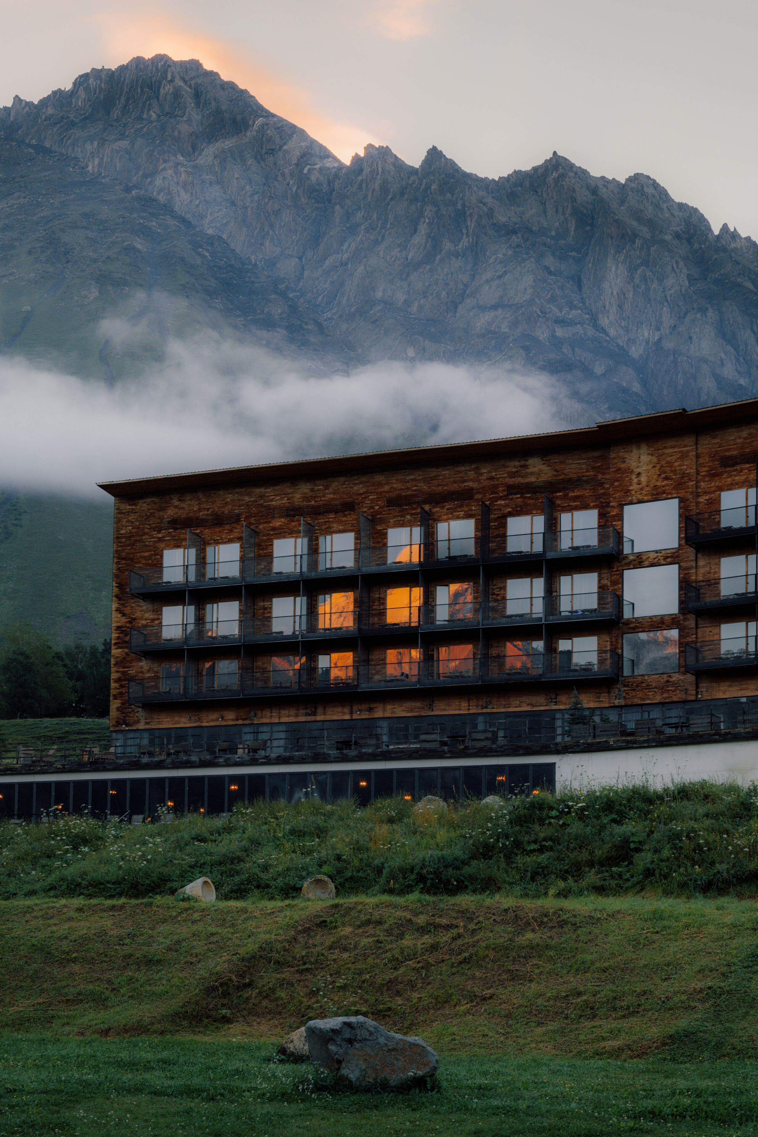 A modern building with multiple windows and a wooden facade, situated in a natural landscape with mountains in the background.