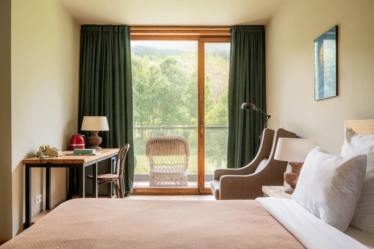 Quarto de hotel com cama, escrivaninha com luminária, cadeira e vista para uma floresta através de uma grande janela.
