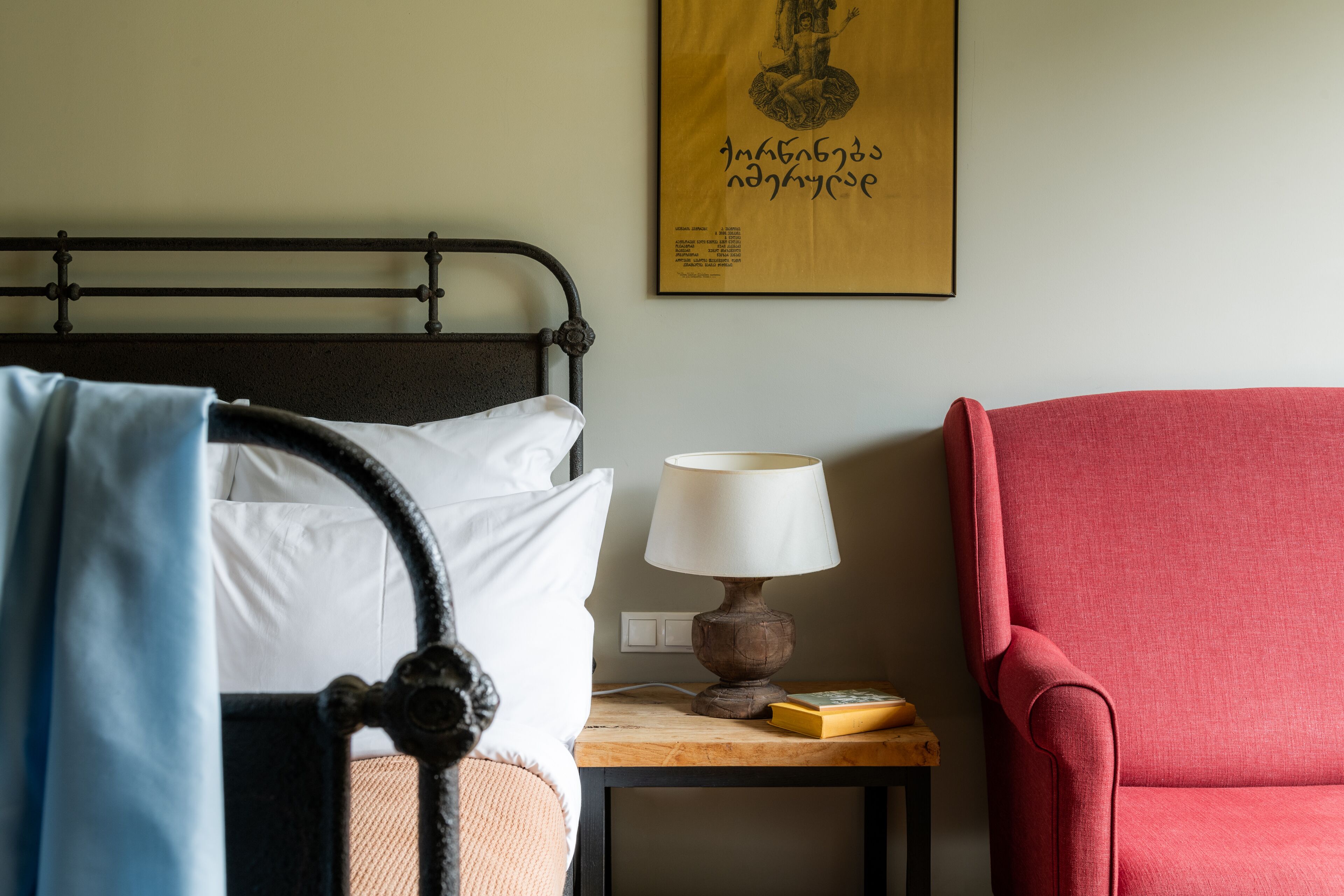 Una cama bien hecha con sábanas blancas, una mesita de noche de madera con una lámpara y un libro, y un sillón rojo.
