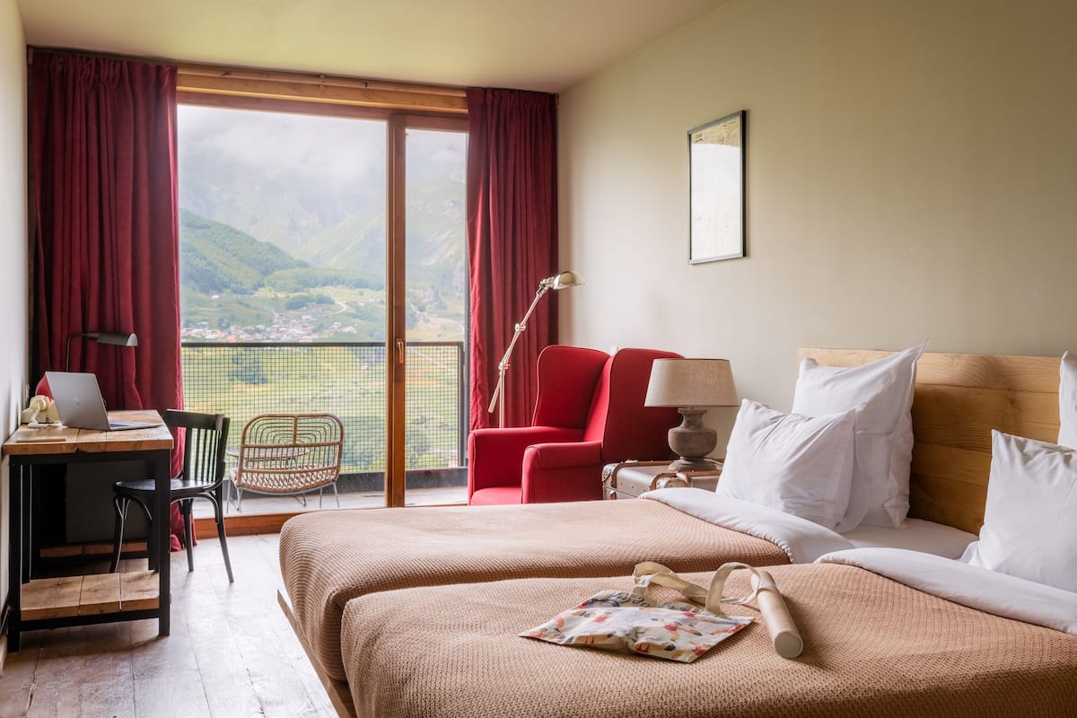 Quarto de hotel com cama, escrivaninha com laptop, cadeira, luminária e vista para montanhas através de amplas janelas.