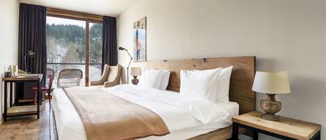 A hotel room with a wooden bed, a desk, two chairs, a nightstand with a lamp, and a window with a view of trees.