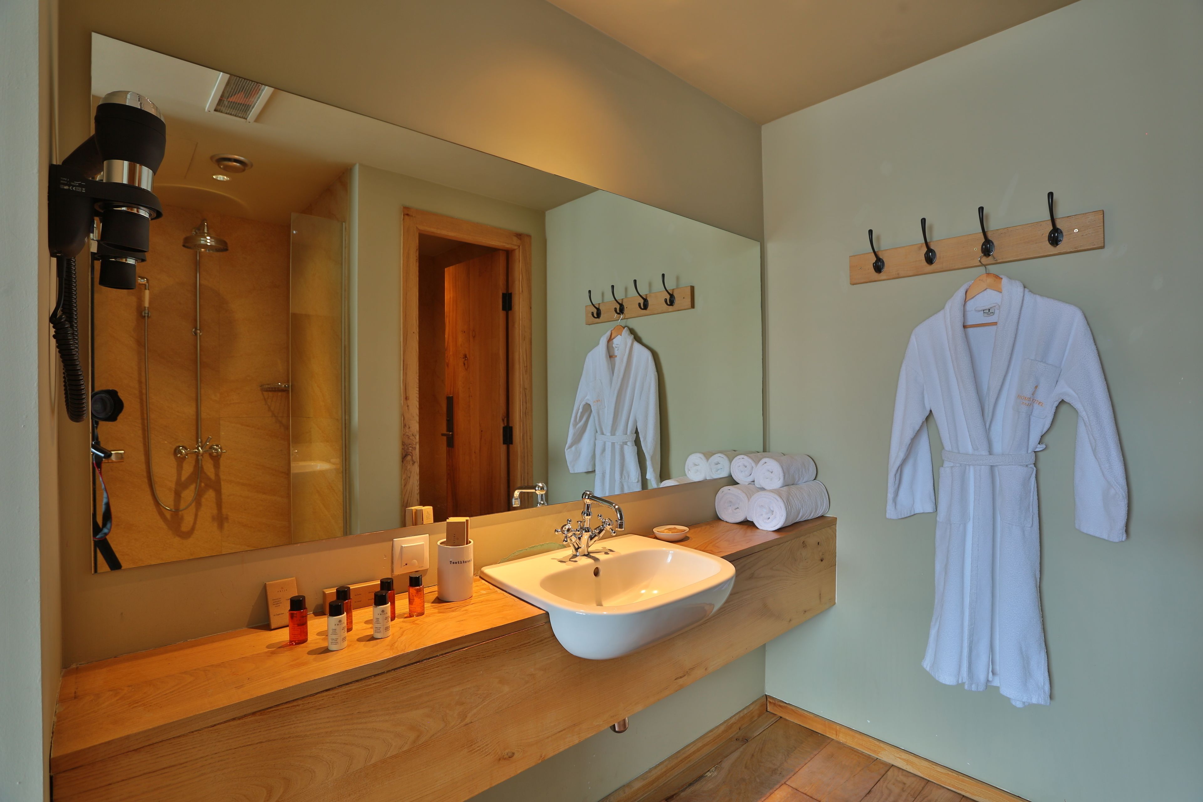 A bathroom with a wooden vanity, a white sink, a large mirror, a hairdryer, and a towel rack with two white robes.