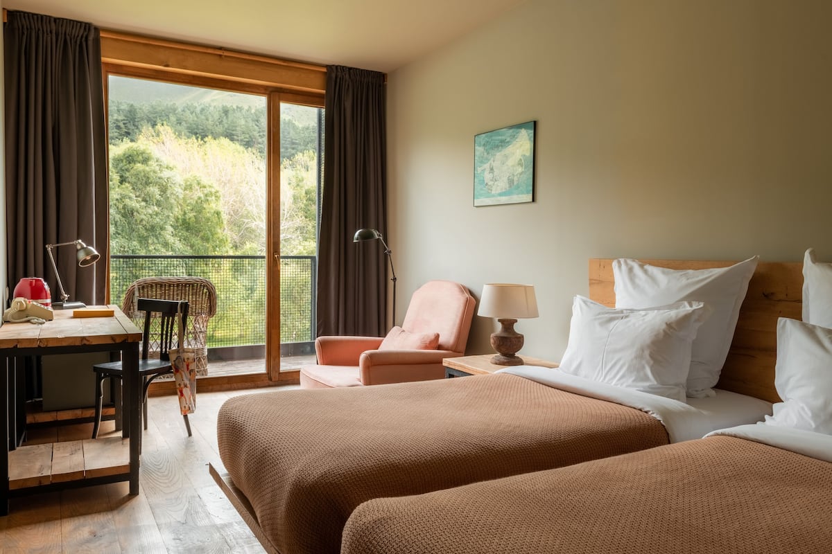 Quarto de hotel com duas camas, uma mesa, uma cadeira e vista para uma floresta por uma grande janela.
