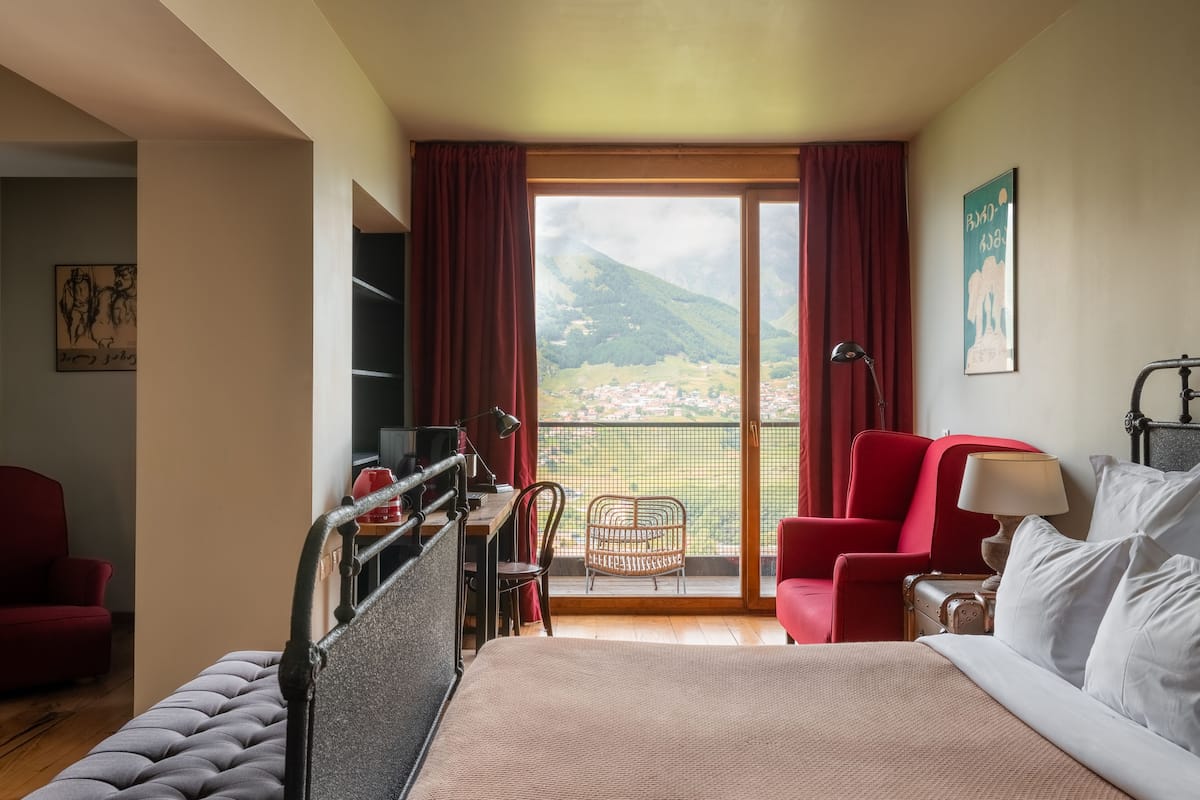 Quarto de hotel com cama, escrivaninha, cadeira e varanda com vista para uma cidade e montanhas.