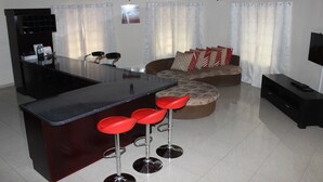 Une cuisine moderne dotée d’un îlot central et de tabourets de bar, un espace de vie avec un canapé et des coussins, et un téléviseur à écran plat.